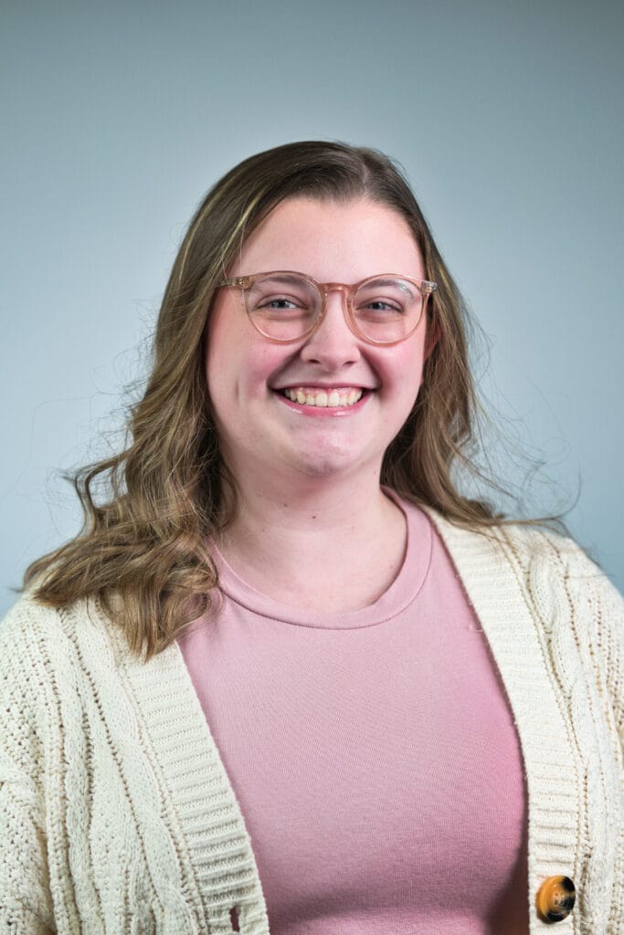 A young woman with wavy light brown hair wearing glasses, a pink shirt, and a cream-colored knit cardigan smiles in front of a plain light gray background.